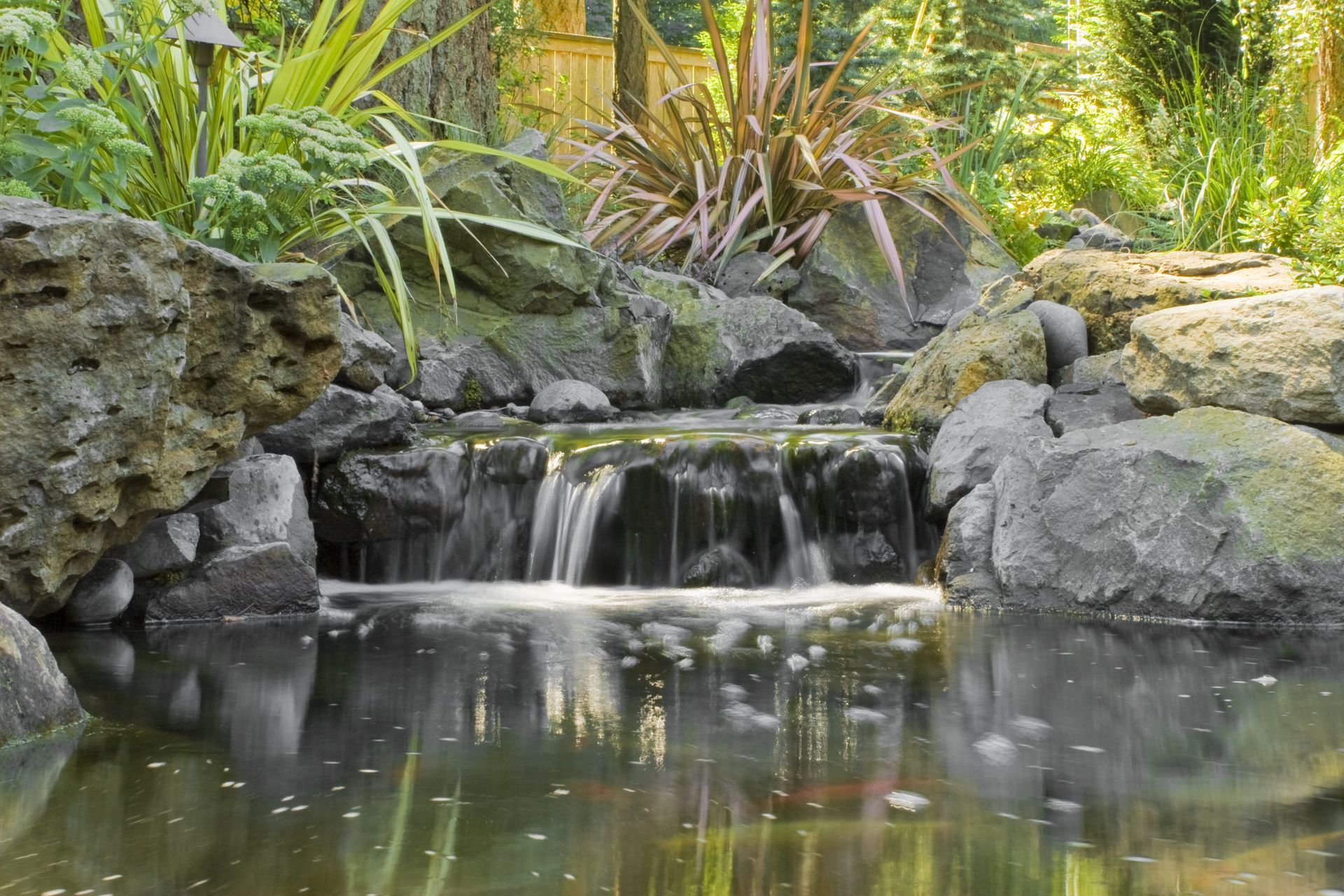 Small garden waterfall cascading over rocks into a calm pond, surrounded by lush green plants.
