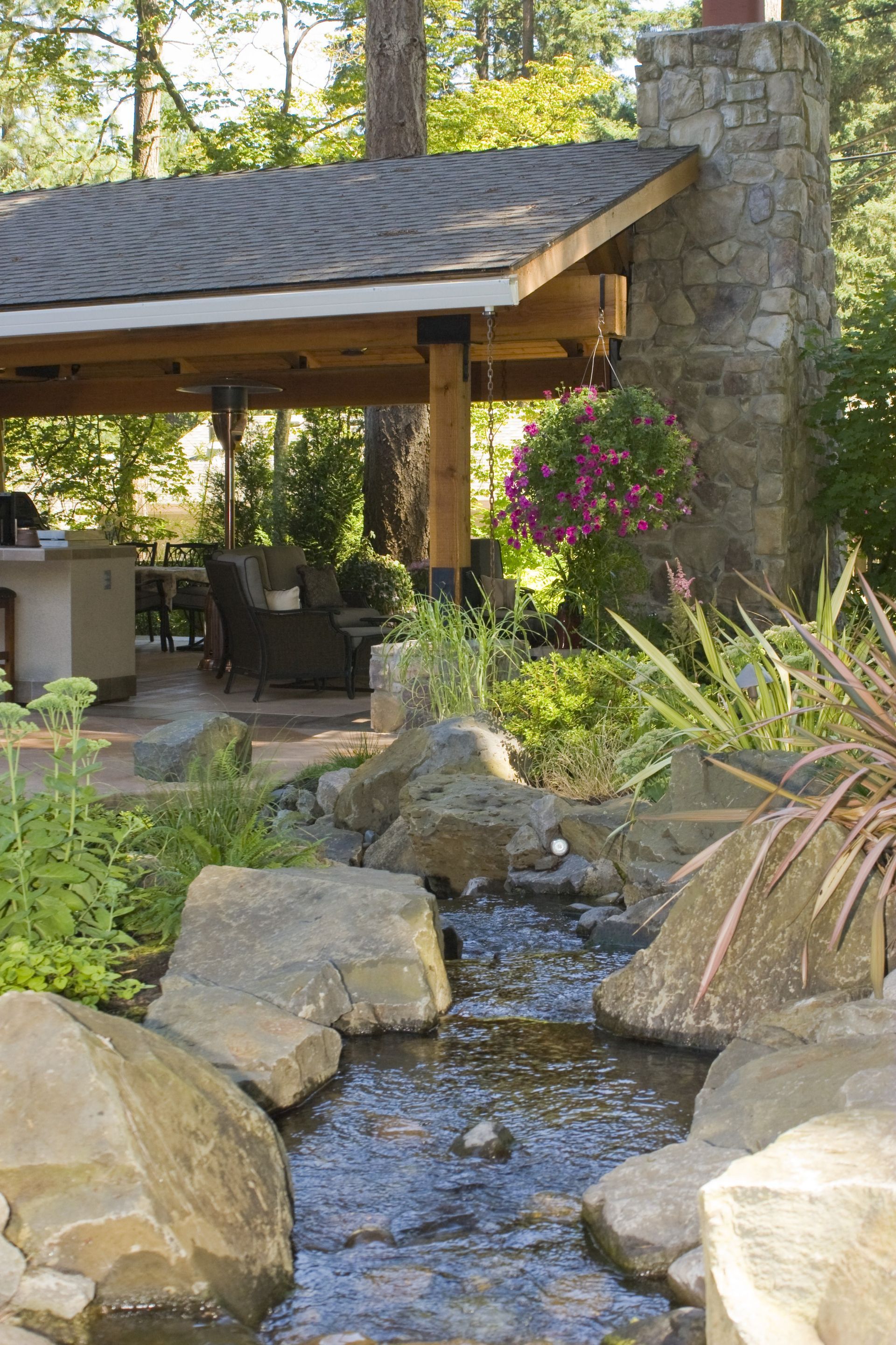 Stone garden stream beside a covered patio and fireplace in a wooded backyard