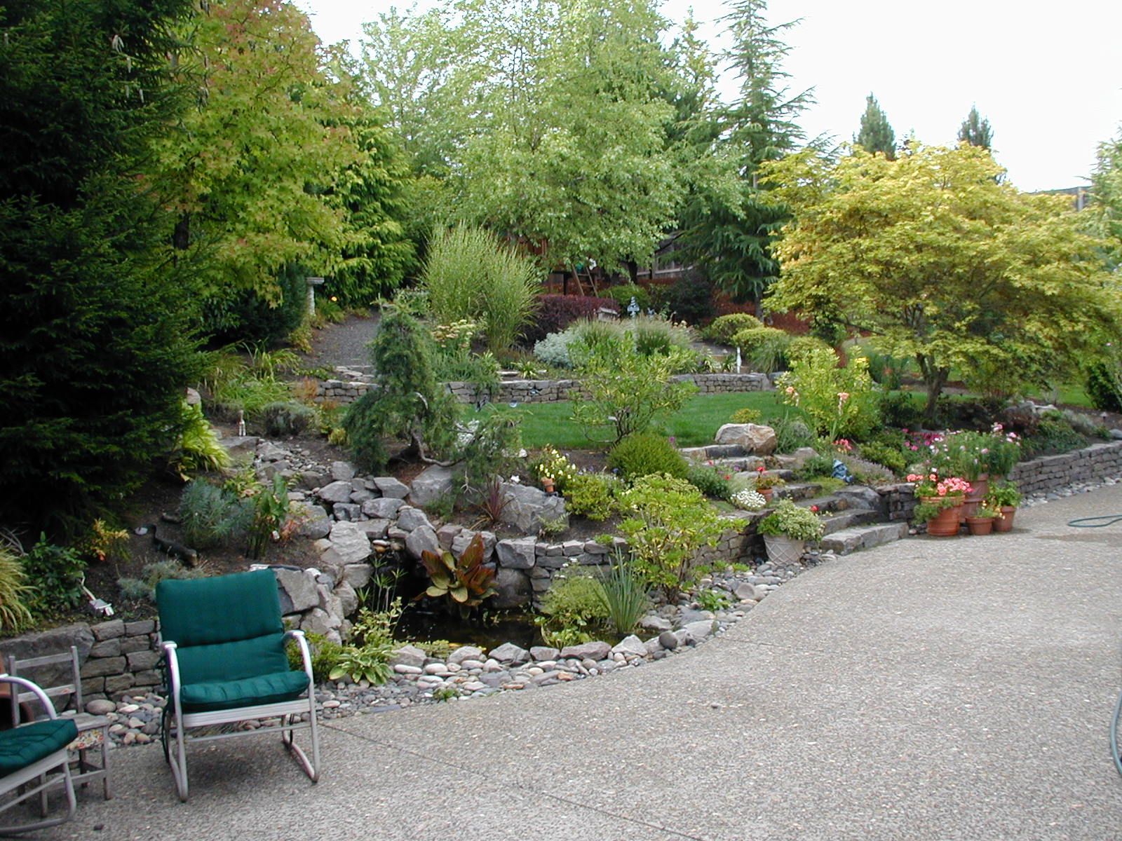 Rock garden landscaping beside a gravel path, with green trees, shrubs, and teal chairs in the foreground