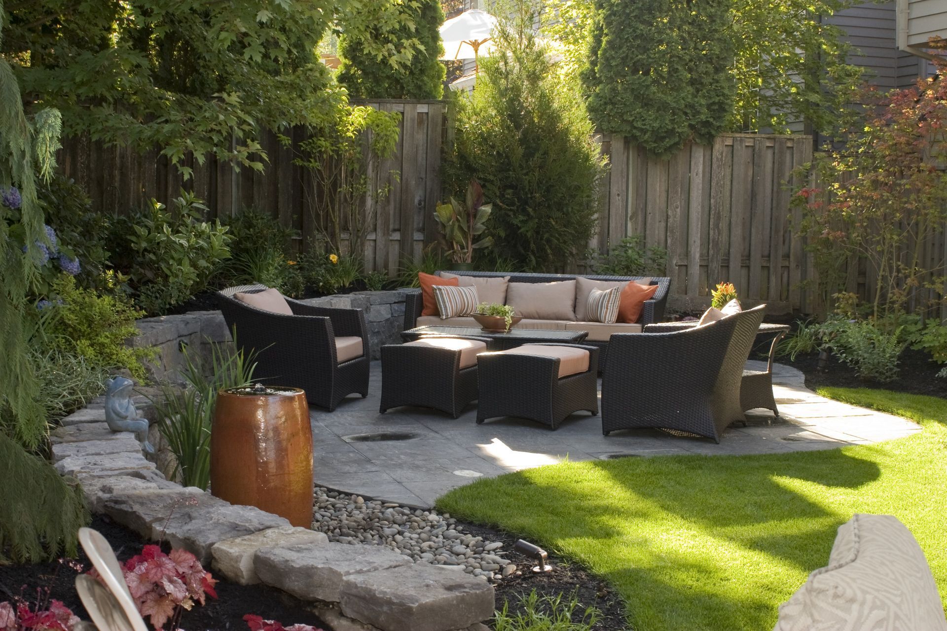 Backyard patio with wicker seating, stone fire pit, and lush green lawn surrounded by trees and fence