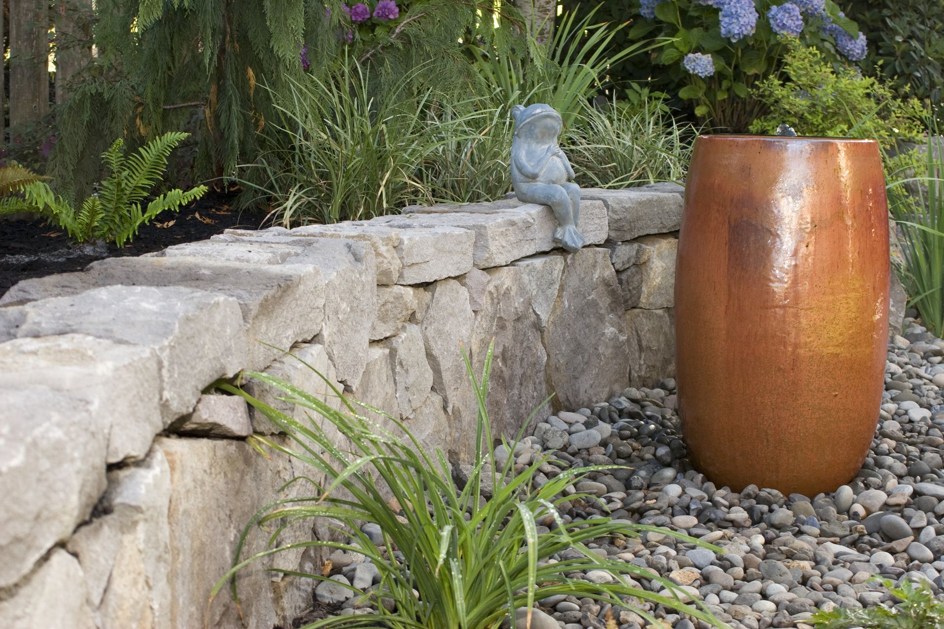 Stone garden wall with small statue beside a tall copper planter on a pebble bed