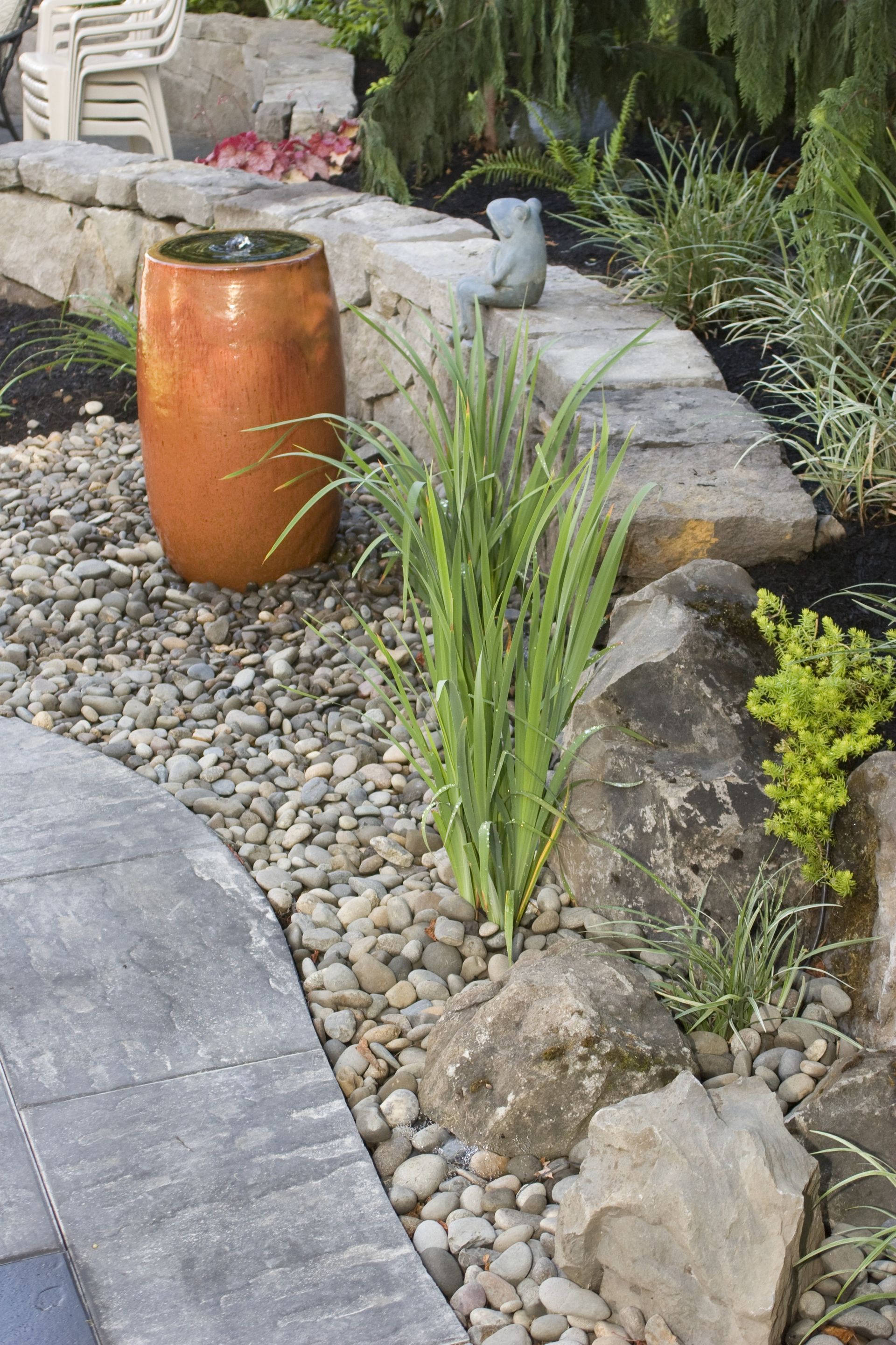 Landscaped garden path with gray pavers, pebbles, rocks, ornamental grass, and a copper rain barrel