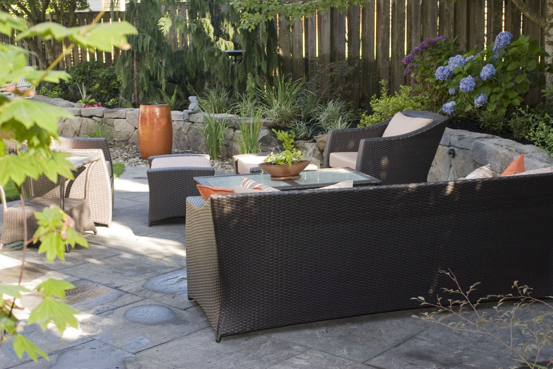 Patio with dark wicker outdoor sofa, chair, table, and potted plants in a sunny garden.