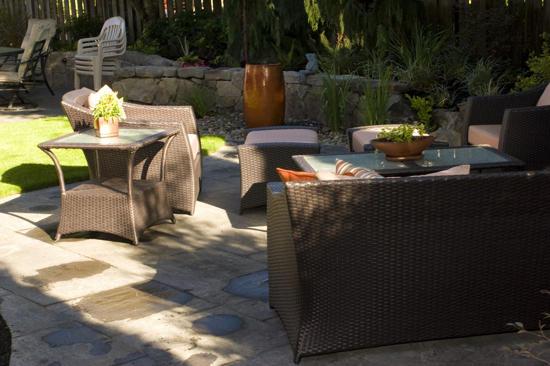 Patio seating area with wicker chairs, glass tables, and potted plants in sunlight
