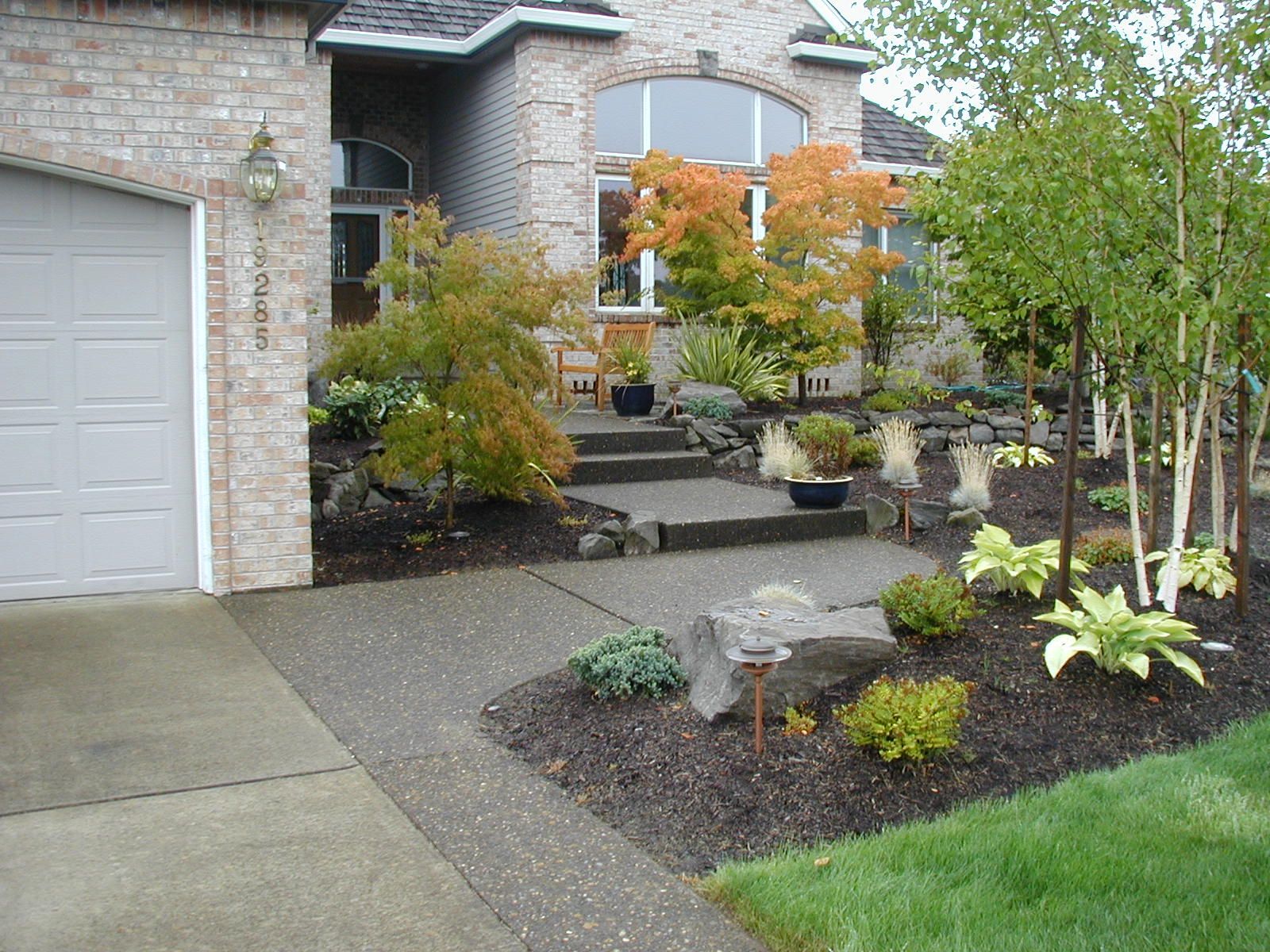 Front yard landscaping with stone steps, shrubs, and a garage beside a brick house