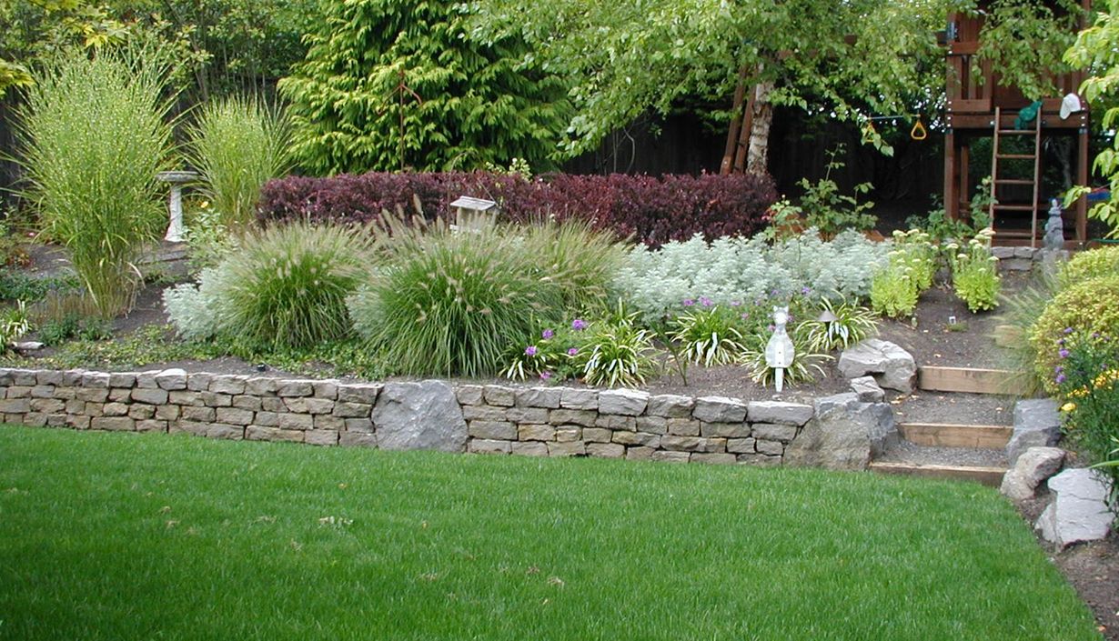 Stone garden wall with lush green lawn, ornamental grasses, and flowering plants in a landscaped yard