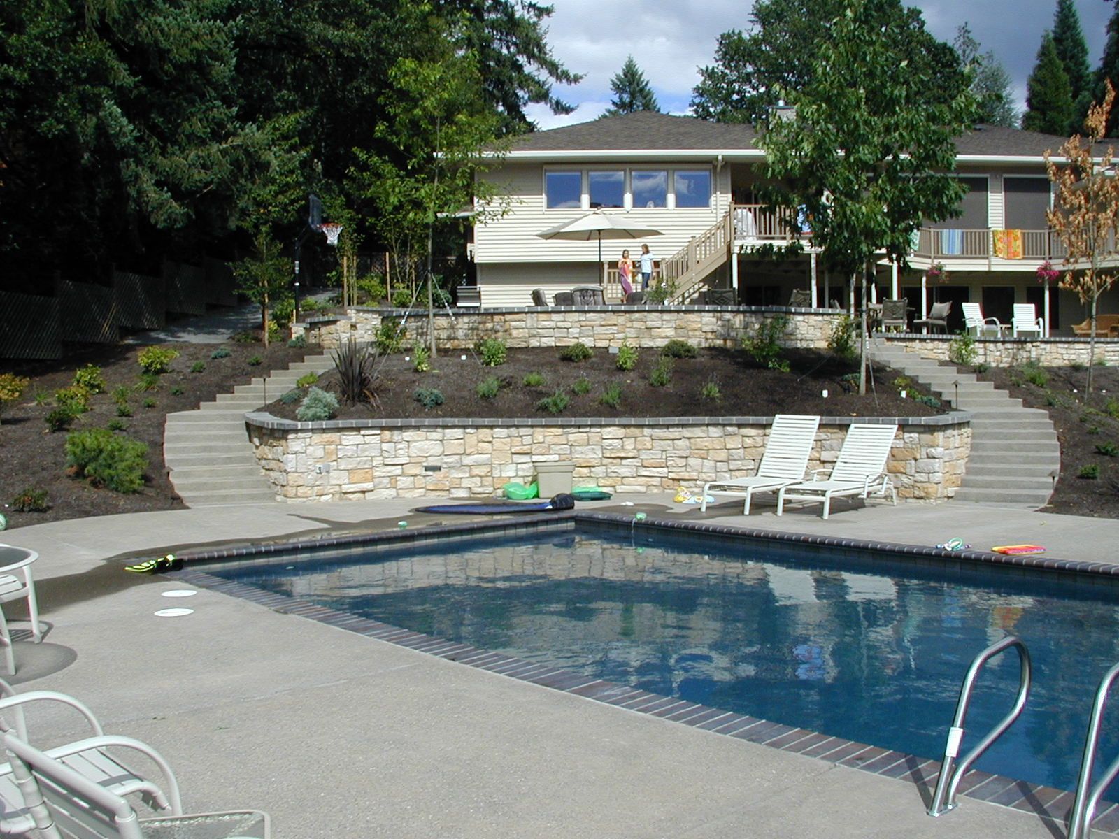 Backyard swimming pool with lounge chairs, stone patio, and houses in the background