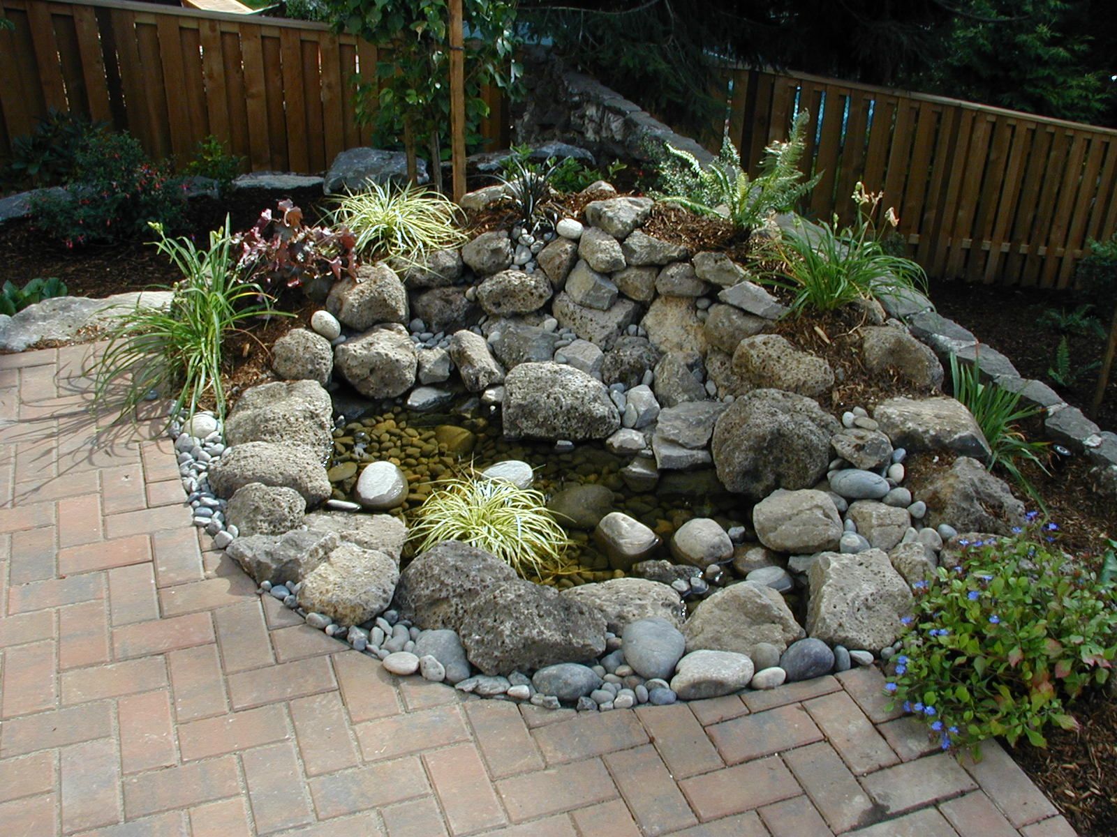 Rock garden with stacked stones, small pond, and potted flowers beside a brick patio and wooden fence