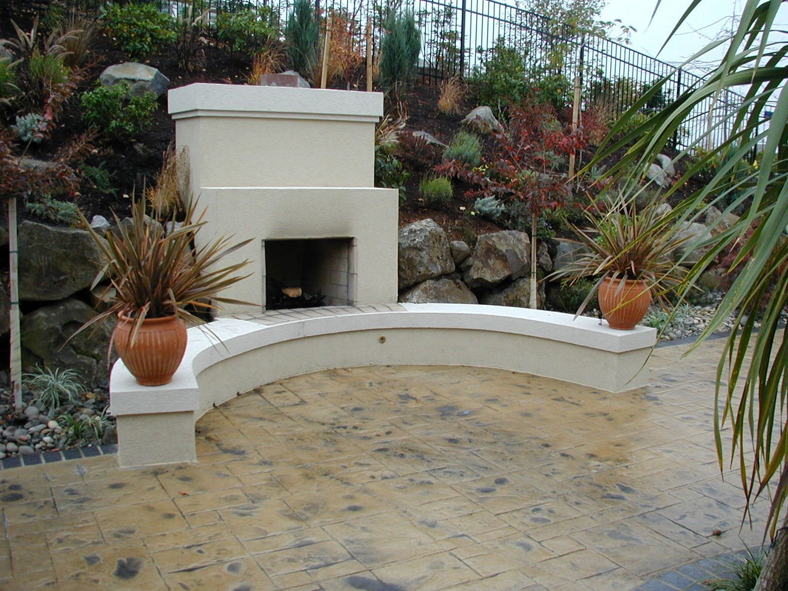 Curved backyard patio with built-in stucco fireplace, potted plants, and stone landscaping