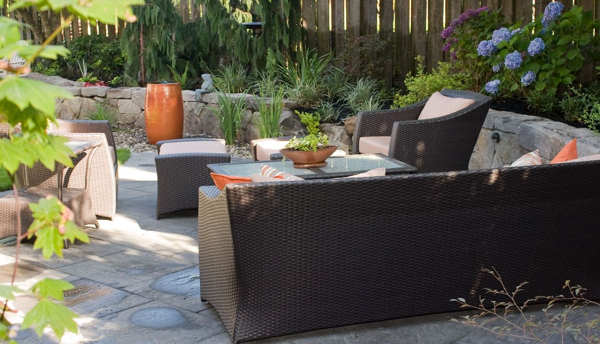 Backyard patio with wicker furniture, potted plants, and a stone table in a sunny garden