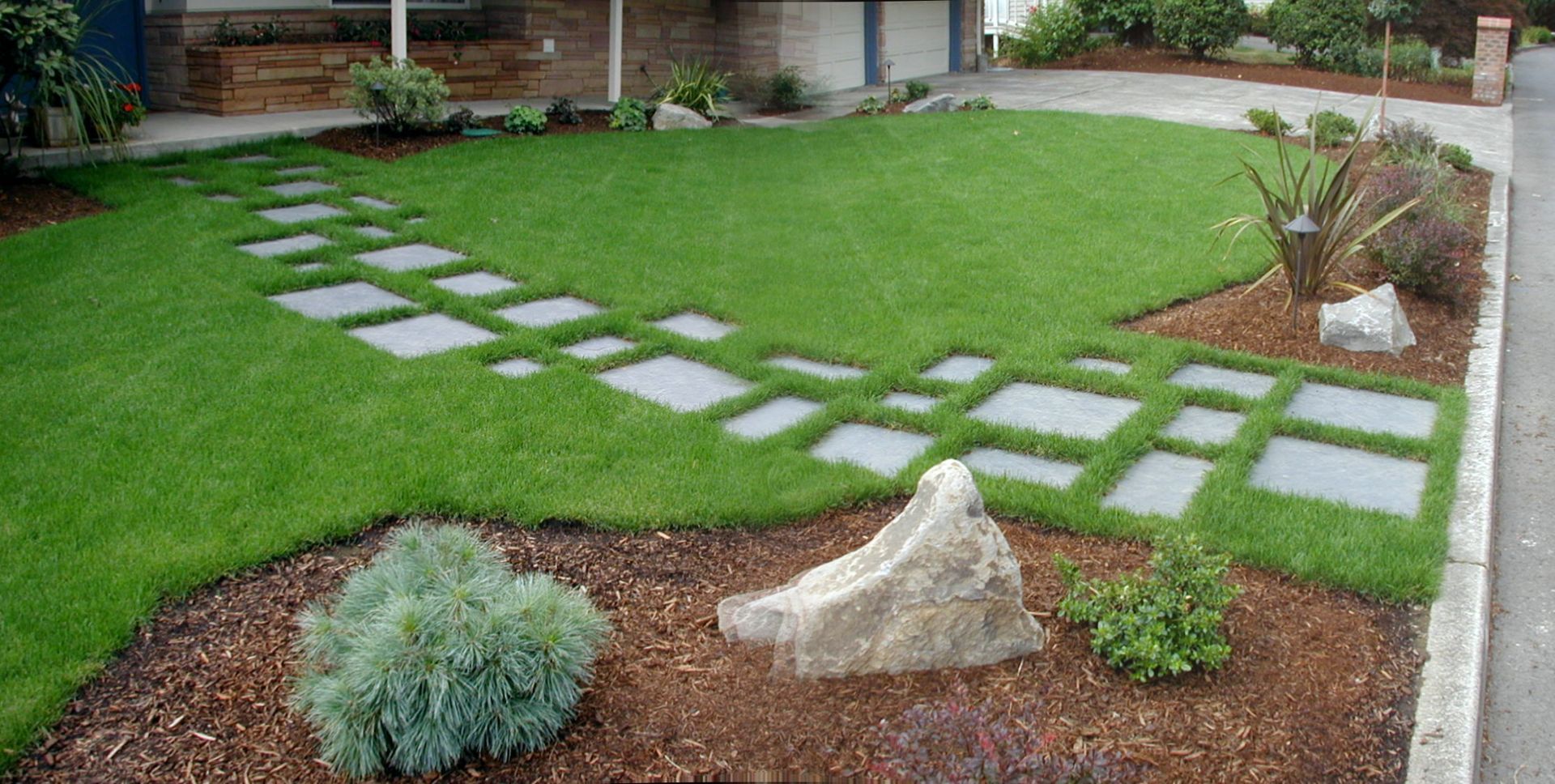 Modern backyard with green lawn, stepping stones, and landscaped mulch beds with shrubs and rocks