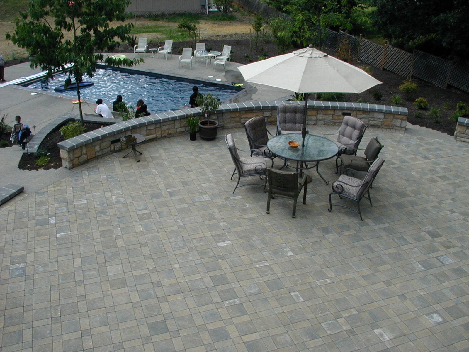 Patio with stone pavers, outdoor table and chairs, white umbrella, and a curved pool with people nearby.