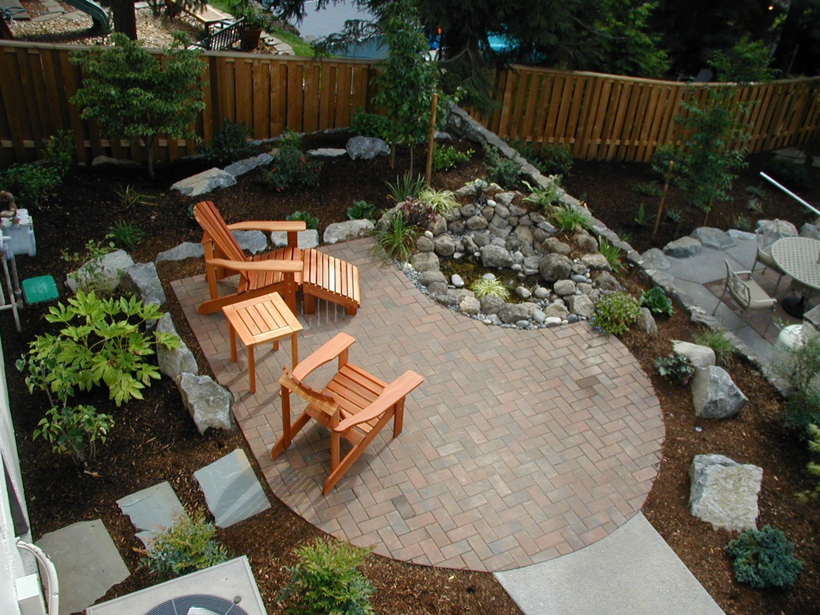 Backyard patio with wooden chairs around a circular brick pad beside a rock garden and fence