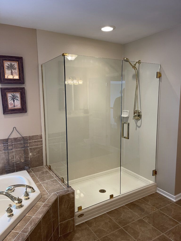 A clear glass corner shower with gold-toned fixtures and a white base, next to a marble-lined bathtub.