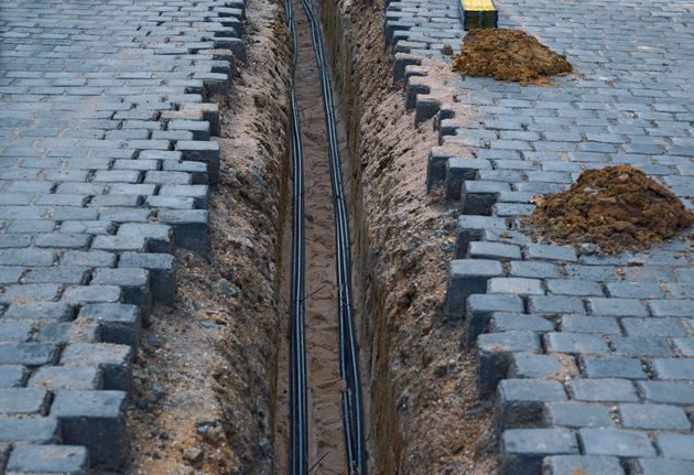 Open trench in gray cobblestone street with exposed underground cables and dirt piles on both sides
