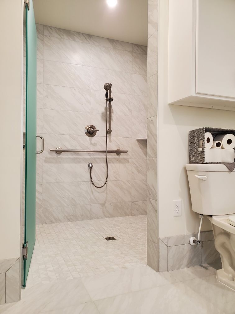 Wheelchair-accessible bathroom with a walk-in shower and toilet. White marble tile, glass door, grab bar.