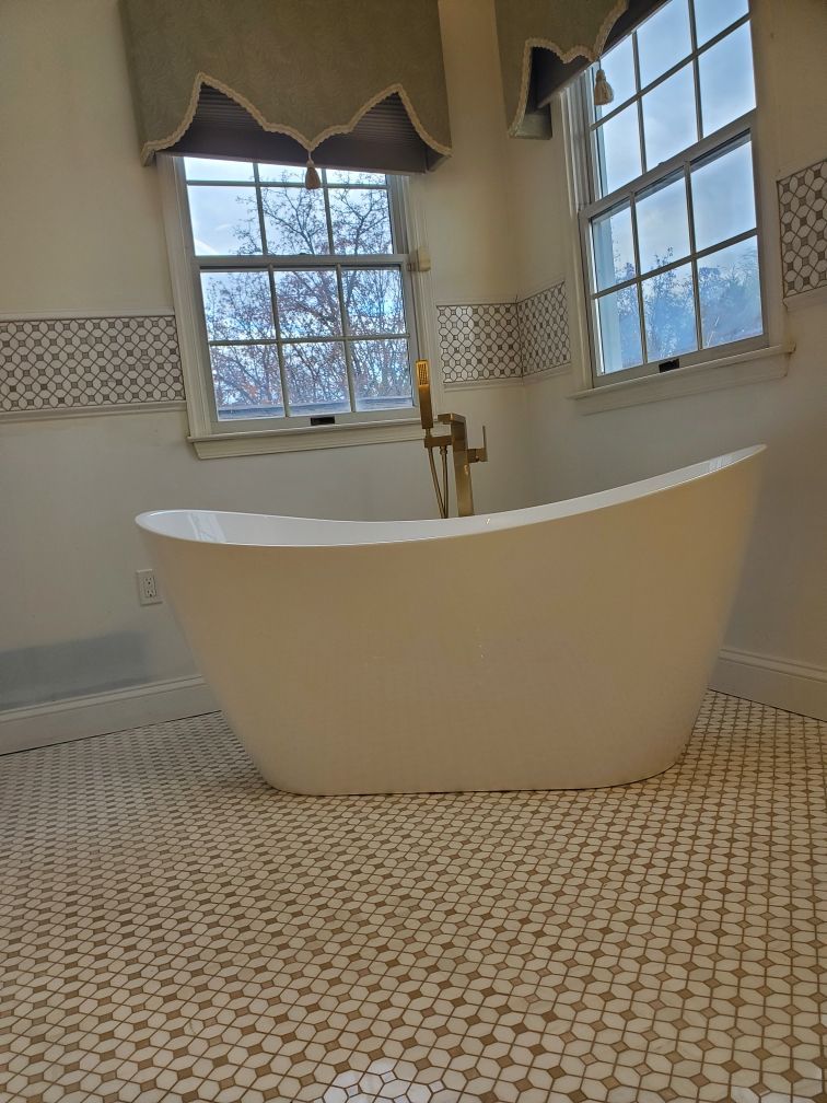 Freestanding white bathtub in a bathroom with mosaic tile floor, two windows, and gold fixtures.