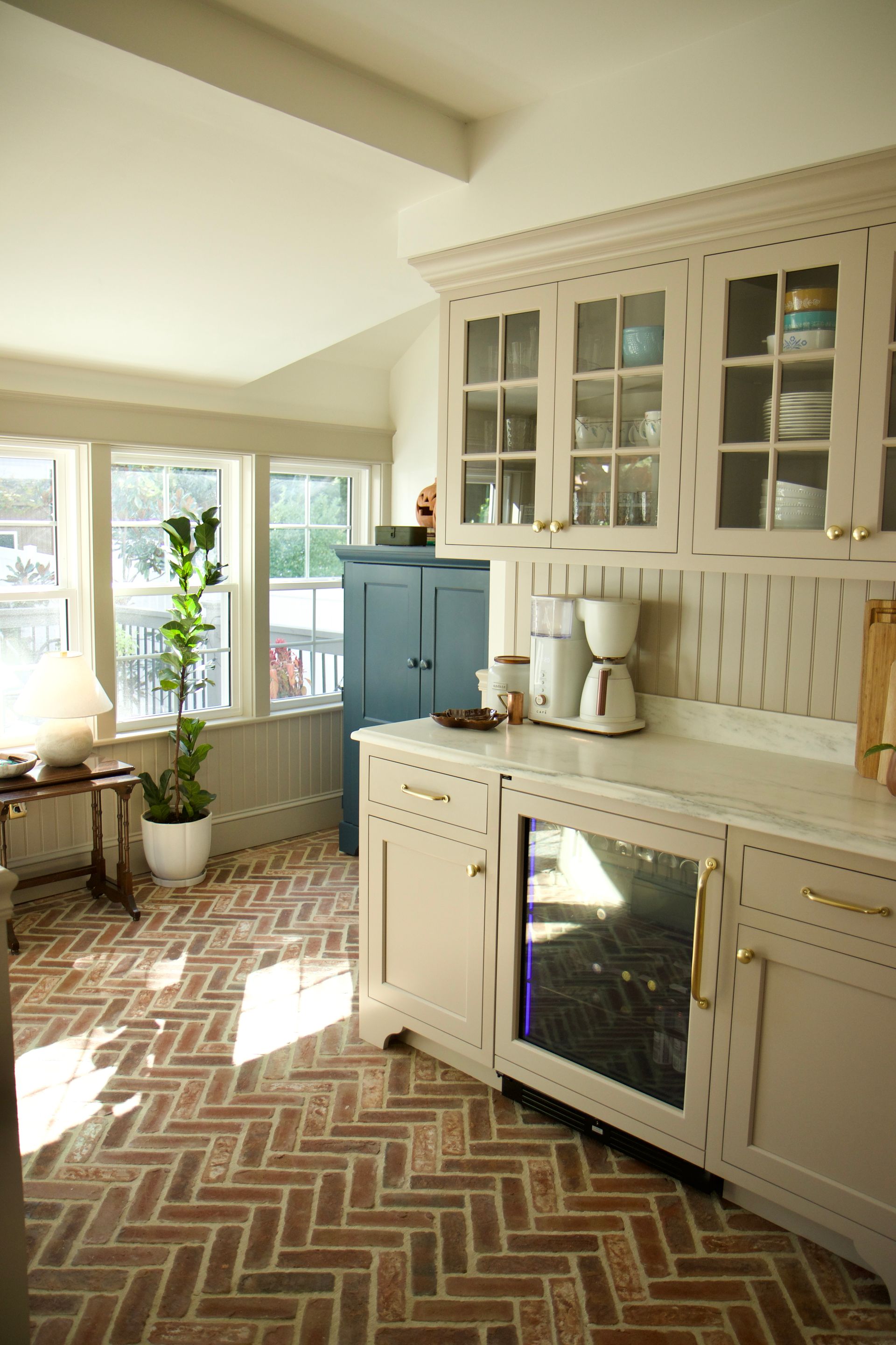 Kitchen with beige cabinets, brick floor, and built-in wine fridge. Sunlight streams through windows.