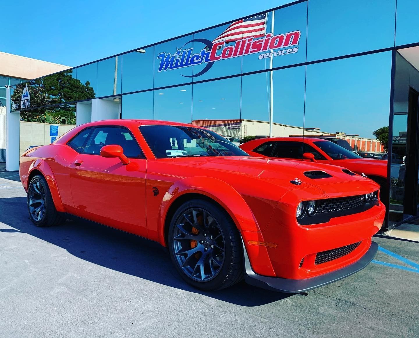 Bright orange Dodge Challenger parked outside a collision repair shop with reflective glass windows.