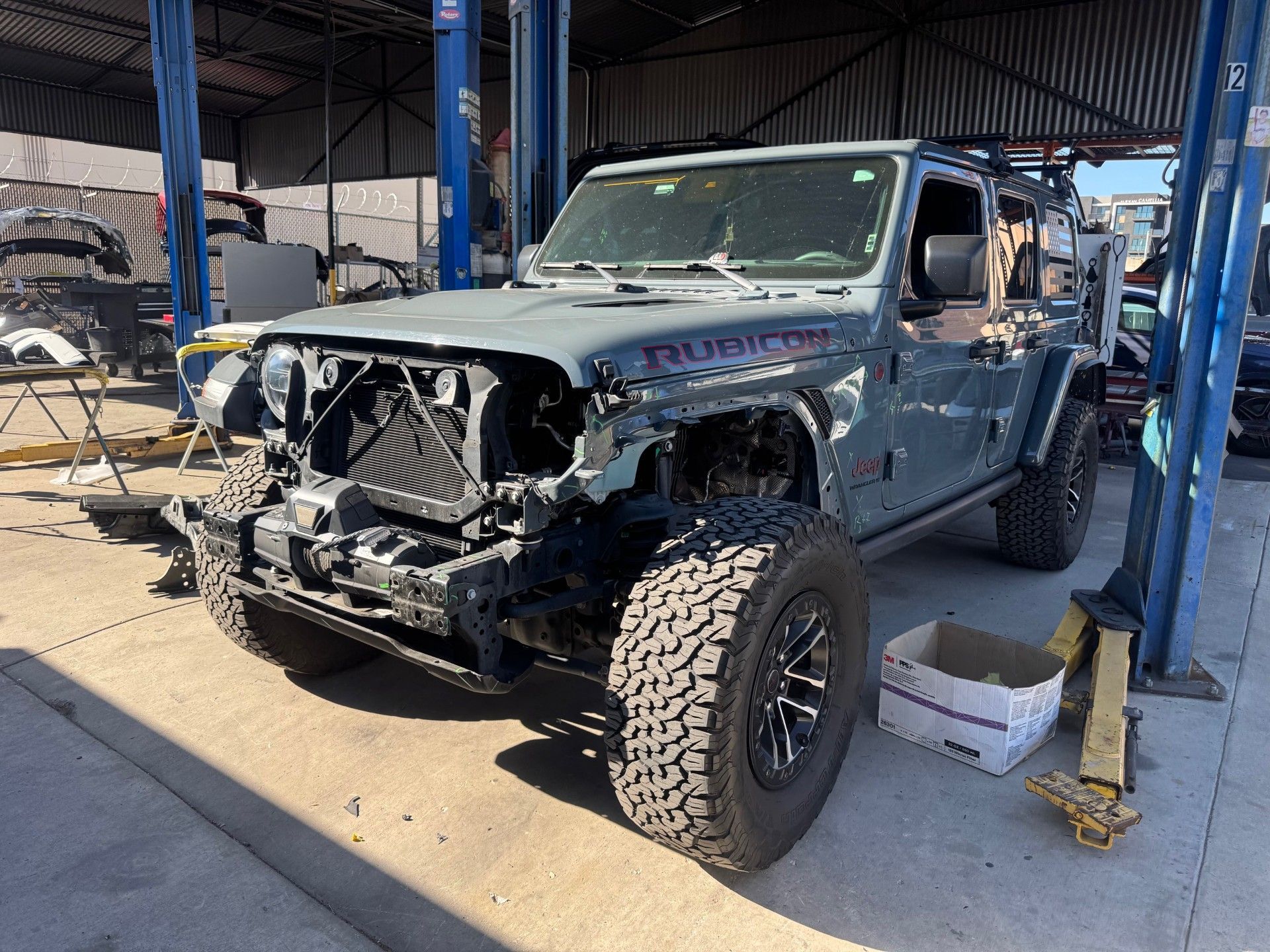 A gray Jeep Rubicon in a repair shop with its front bumper and headlight assembly removed.