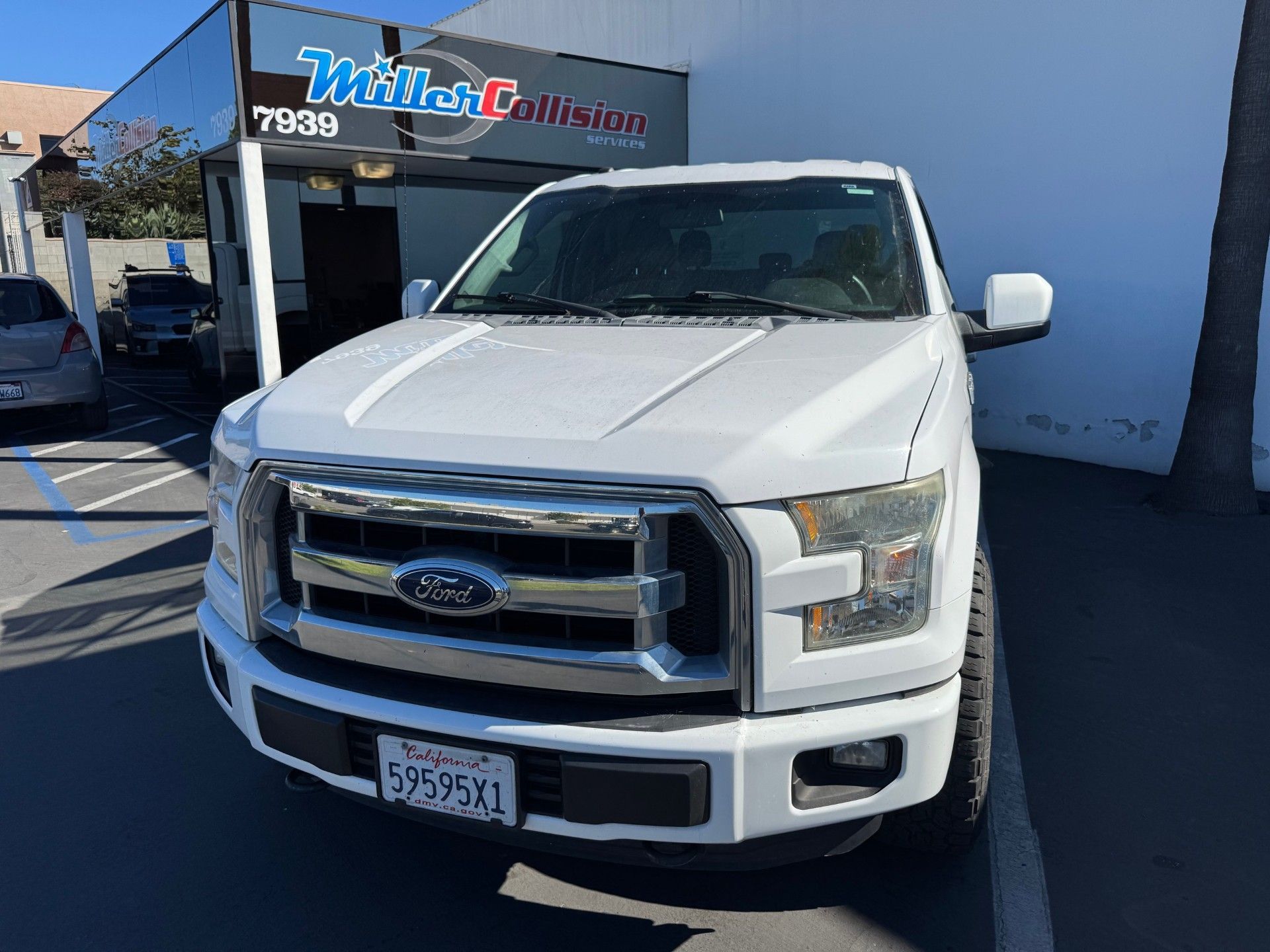 A white Ford F-150 truck parked in front of a collision repair shop under a bright blue sky.