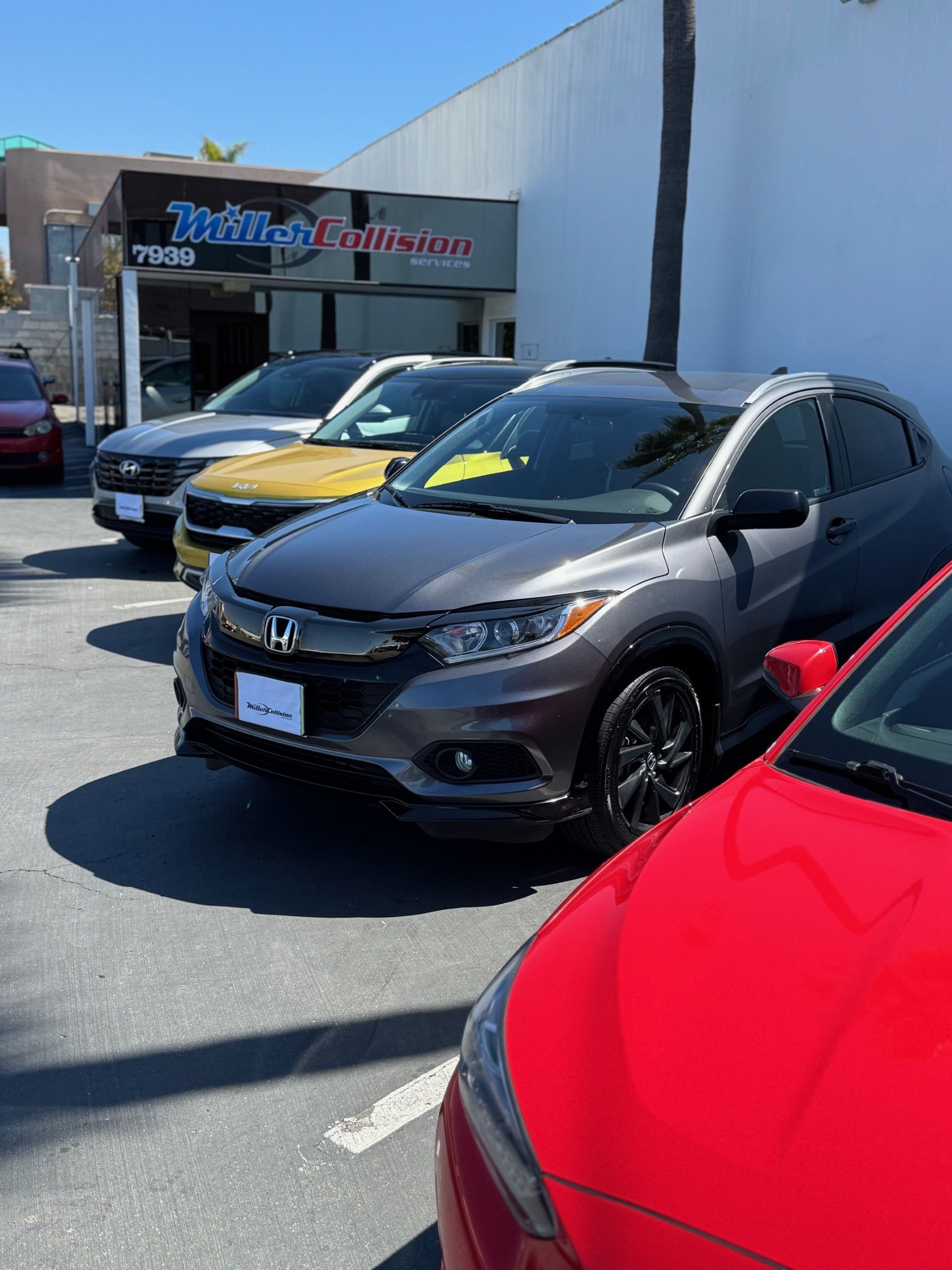 A line of parked cars in front of a dealership building on a sunny day, featuring a grey Honda in the foreground.
