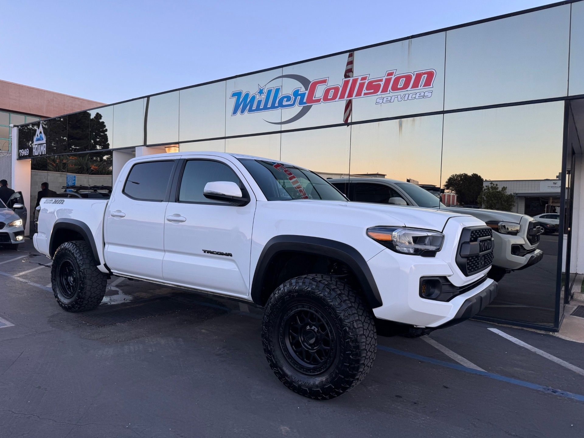 A white Toyota Tacoma pickup truck with black wheels parked in front of a Miller Collision building.