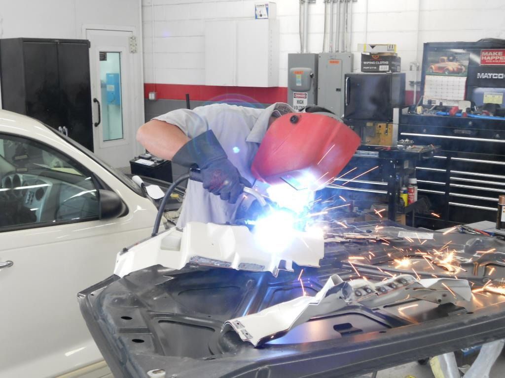 A person welds metal parts, wearing a welding mask and gloves, inside an auto repair shop.