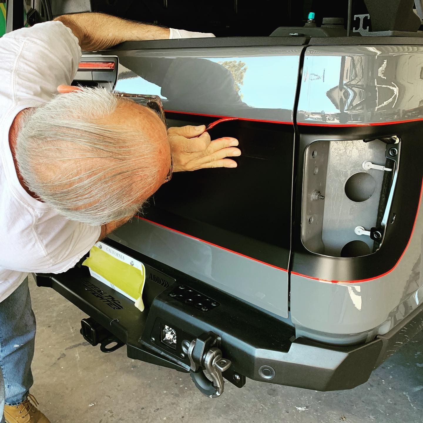 Man installing a black and red trim on a gray truck's bumper and tailgate.