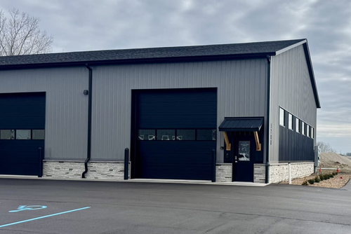 Gray industrial warehouse with black garage doors and a side entrance on an empty parking lot.