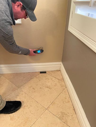 Person using a handheld device to inspect a beige wall corner near a window and tiled floor.