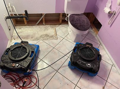 Two black floor machines on blue pads in a tiled room with exposed pipes and a bucket.