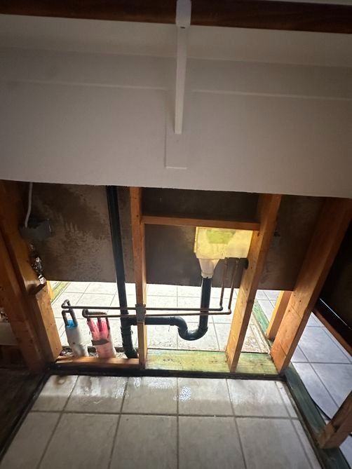 Exposed plumbing and open wall beneath a sink, with tiled floor and tools in a bathroom under repair