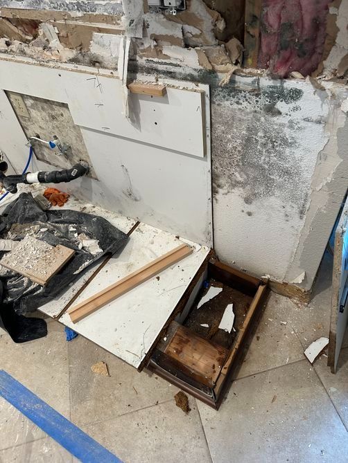 Damaged wall with debris and broken cabinet pieces on a tiled floor, near blue tape marking a work area