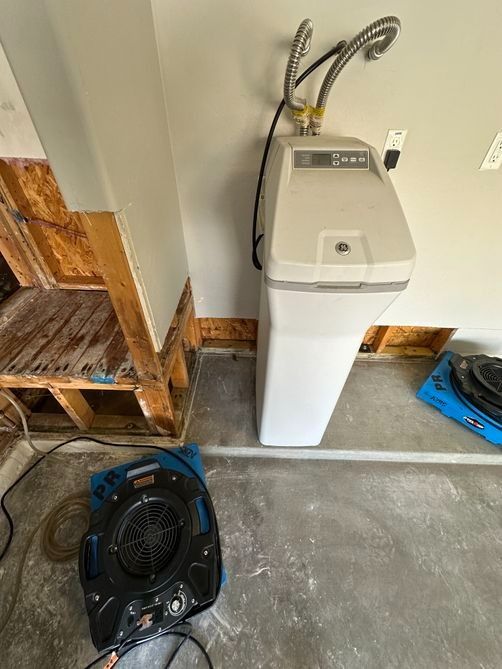 Laundry room with a white washing machine, exposed dryer vent hoses, and a black floor fan on concrete.
