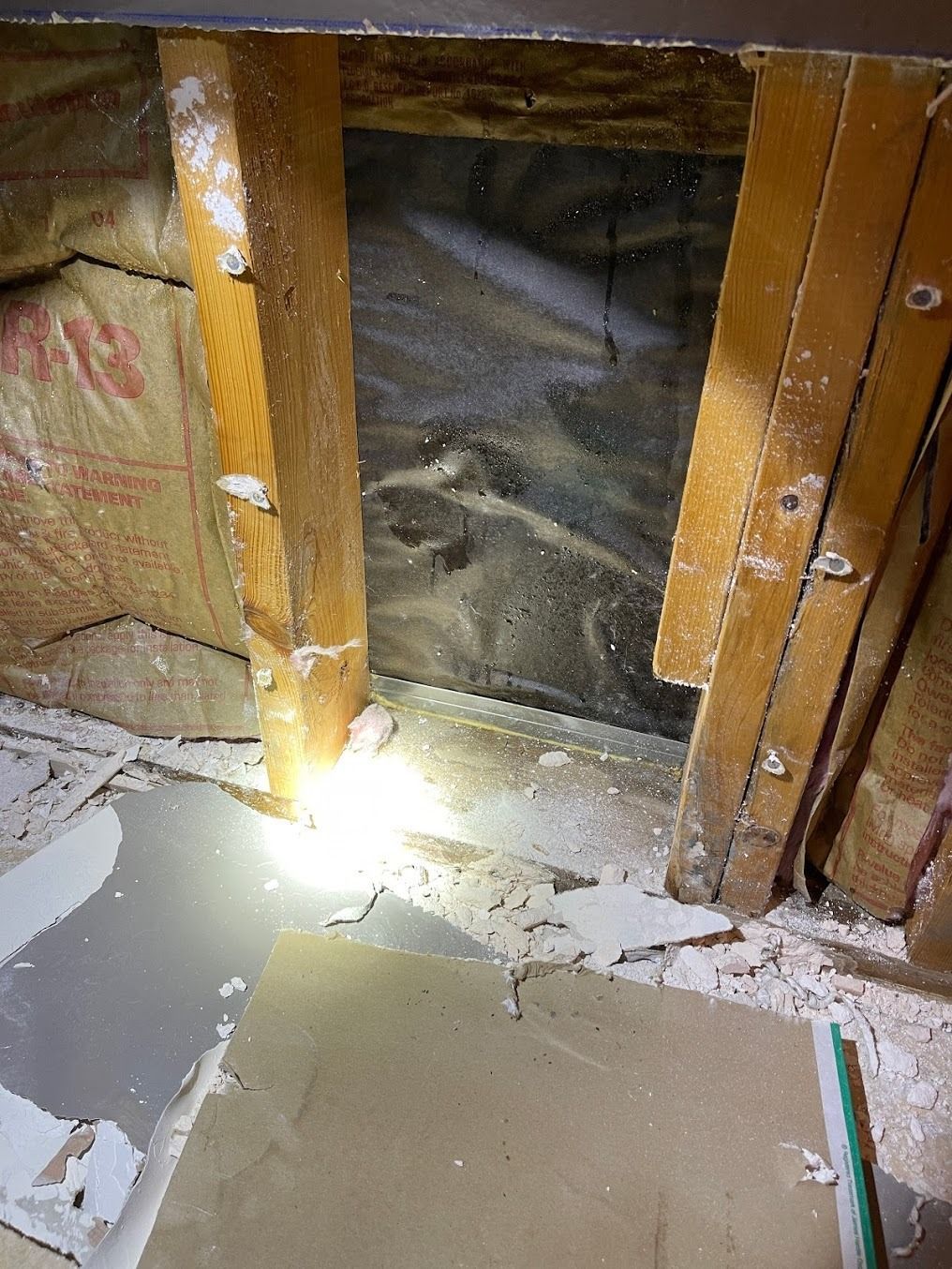 Damaged wall framing with exposed insulation and debris on the floor, lit by a flashlight