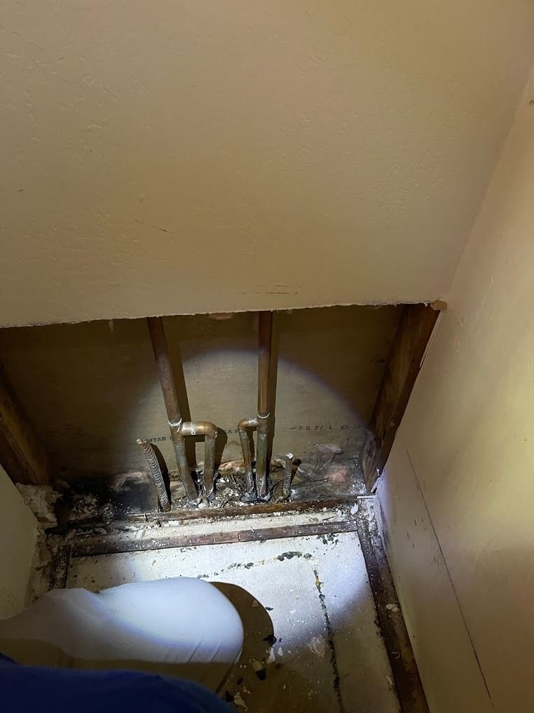 Under-sink plumbing in a damaged wall cavity with exposed pipes and debris on the floor