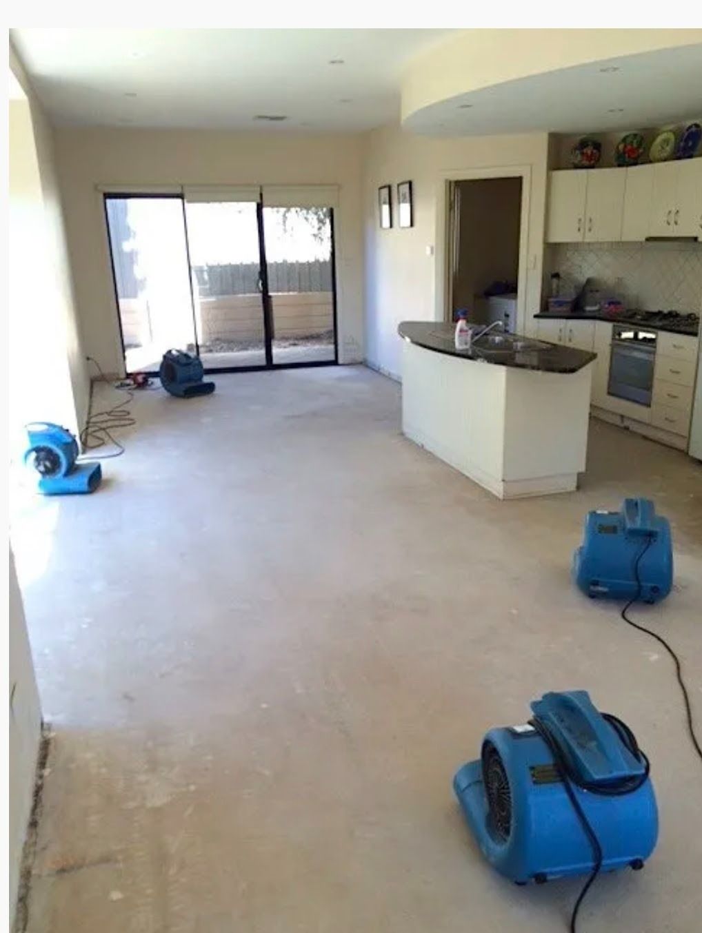 Empty living room with beige carpet, kitchen area, and blue fans on the floor during renovation