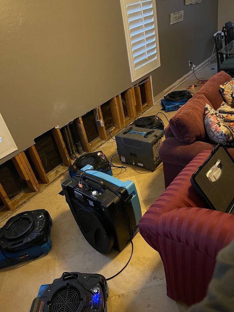 Living room with water-damaged baseboards, exposed wall studs, and several air movers/dehumidifiers running
