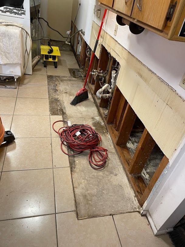 Cluttered kitchen floor with coiled red cord, broom, and cleaning tools beside open cabinet under sink.