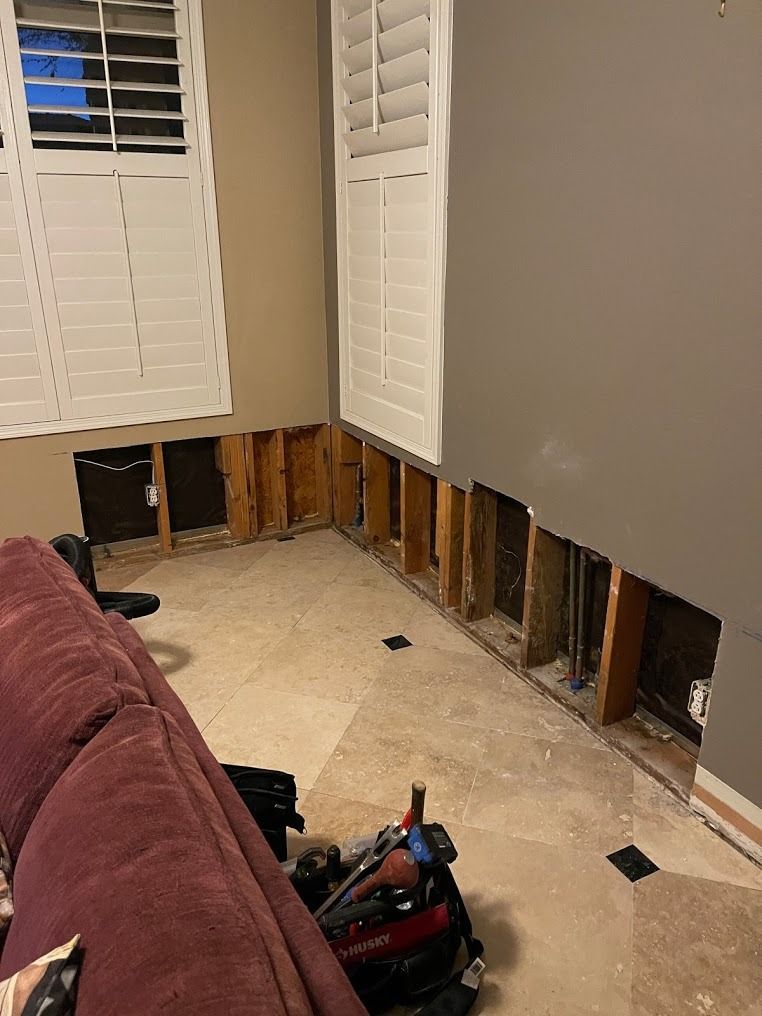 Interior room wall with lower drywall removed, exposing studs beneath white shutters and a maroon couch.