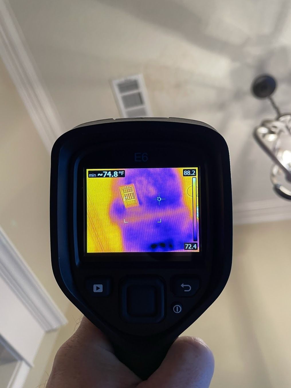 Handheld thermal camera showing a purple heat signature on a ceiling vent indoors