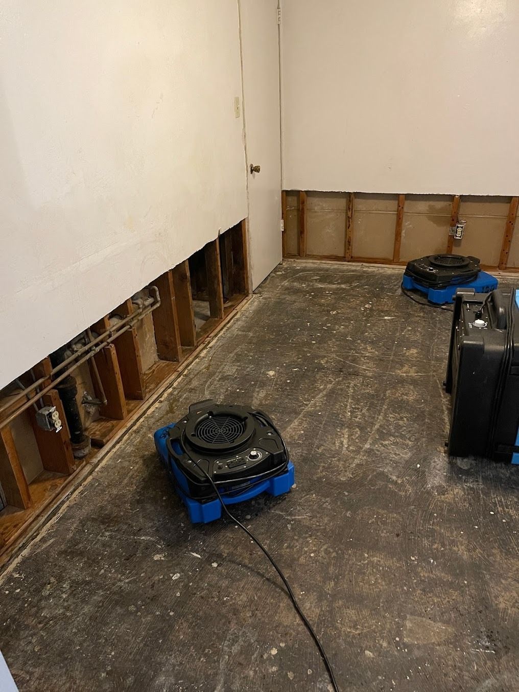 Partially renovated room with exposed wall studs and two blue-and-black floor scrubber machines on a concrete floor