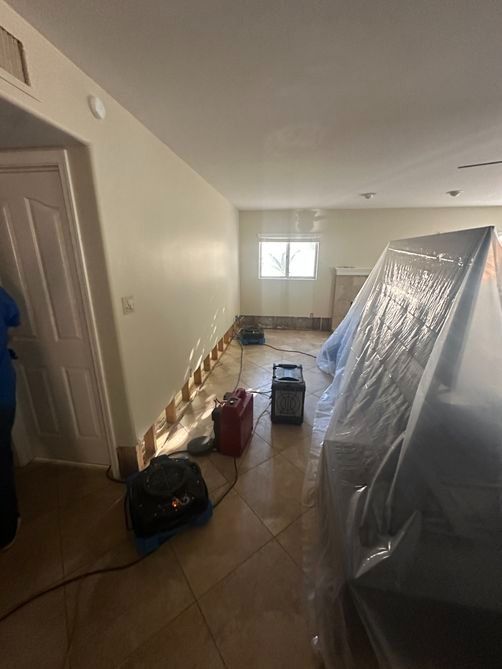 Narrow room under renovation with plastic-covered furniture, tools, and boxes on a tiled floor.