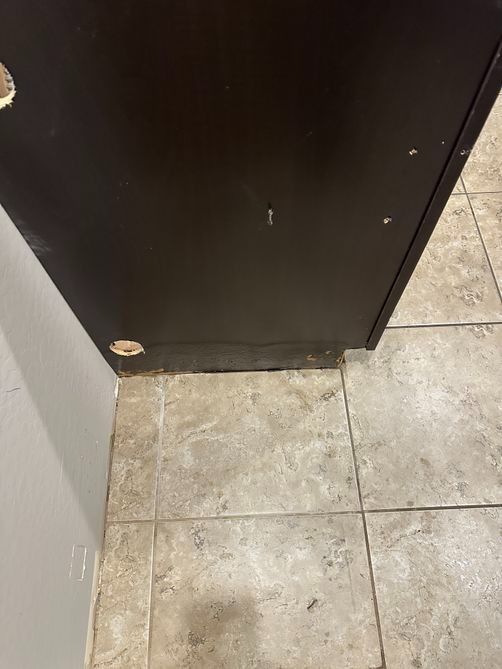 Close-up of a black cabinet corner beside a tiled floor and white wall, with chipped paint at the base.