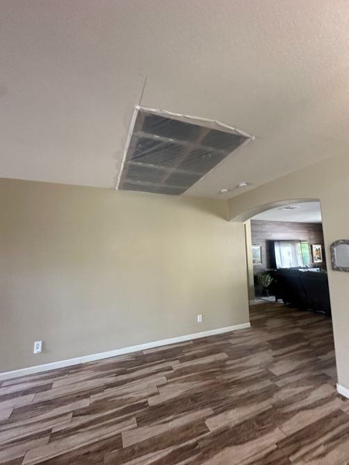 Empty beige living room with damaged ceiling tile and wood-pattern flooring, view into a kitchen area