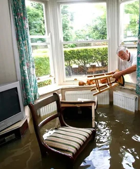 Flooded living room with water up to furniture, and a person carrying a chair by the window.