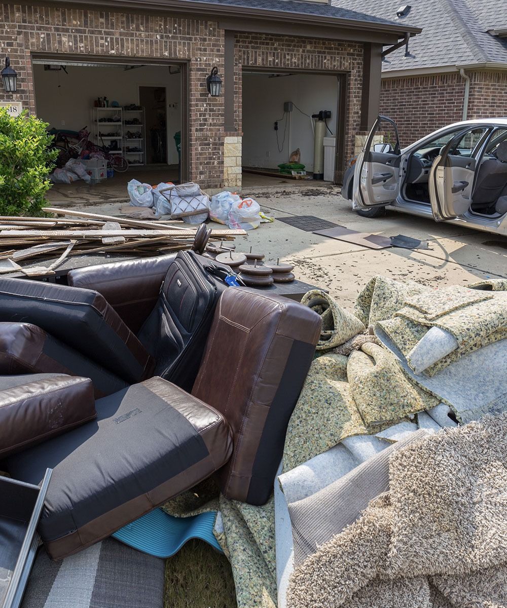 Storm damage in a garage driveway, with scattered furniture, debris, and a damaged car.