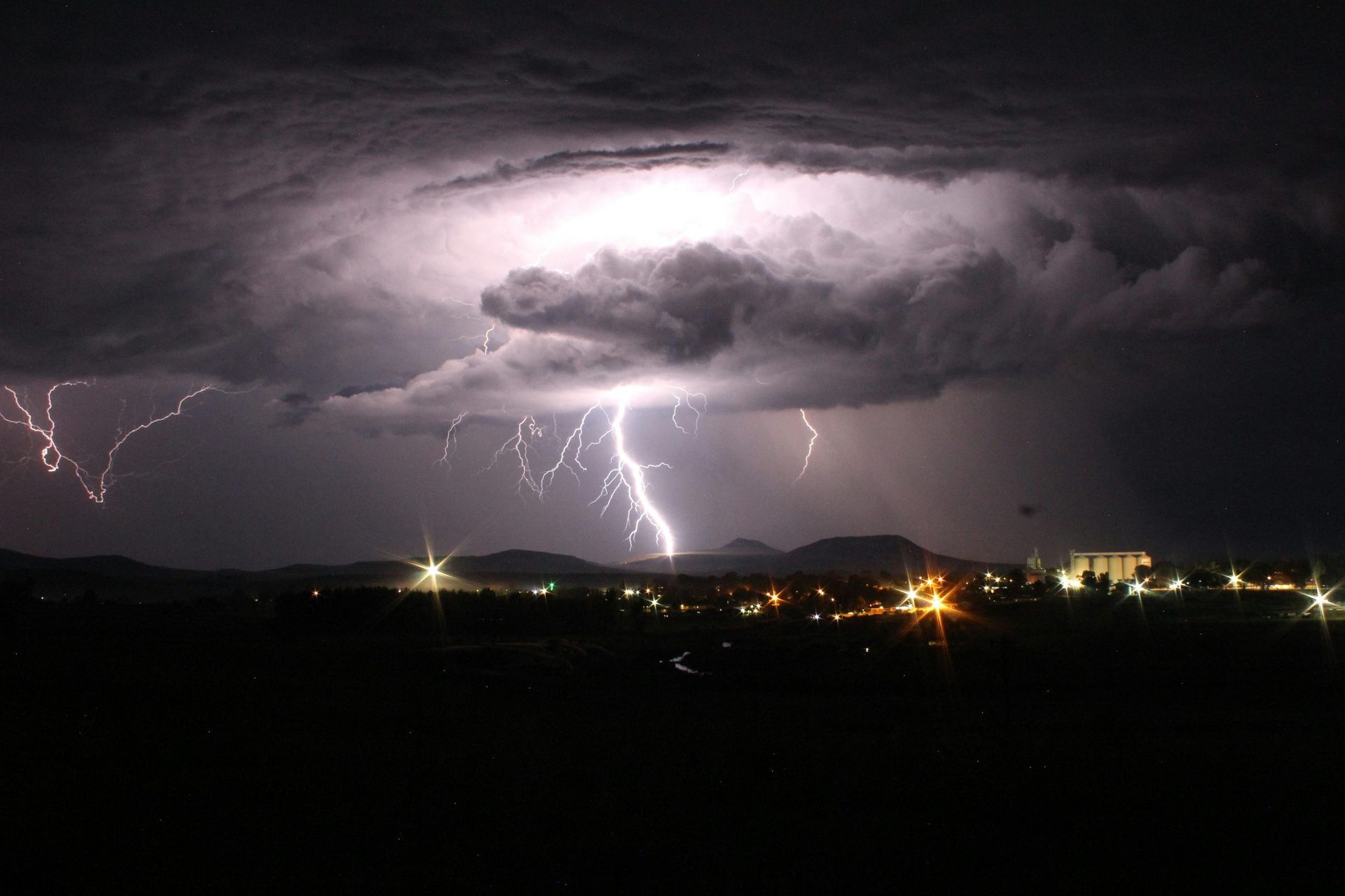 Night lightning storm over a city, with dark clouds and glowing lights on the horizon