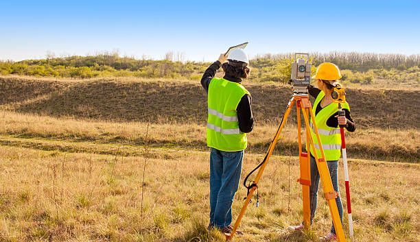 Two surveyors in safety vests using surveying equipment in a field on a sunny day